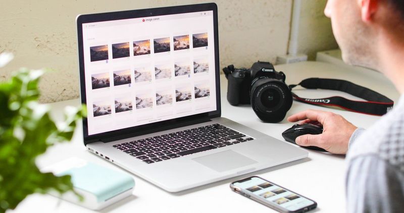 A man looks through images uploaded to image.canon on a laptop with a Canon camera and a Canon SELPHY Square QX10 portable printer on the table alongside. 
