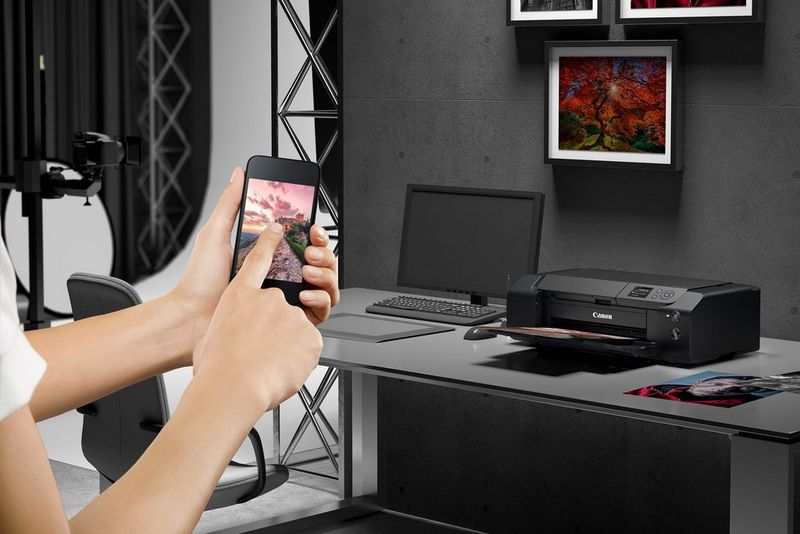 A user taps the screen of a smartphone to print to a Canon imagePROGRAF PRO-300 on the table in front of her. 