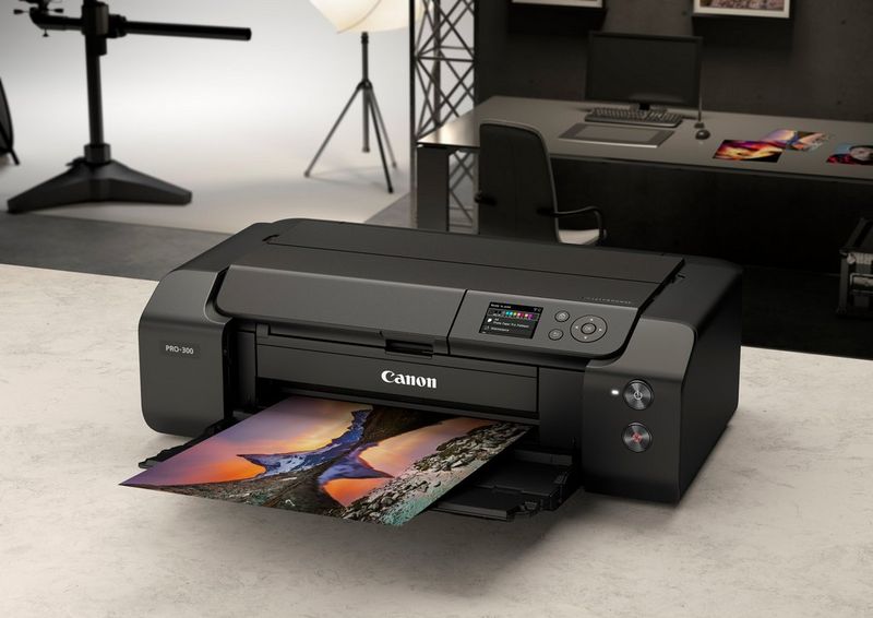 A black Canon imagePROGRAF PRO-300 printer sits on a white desk, a full-bleed colour print resting on its output tray.
