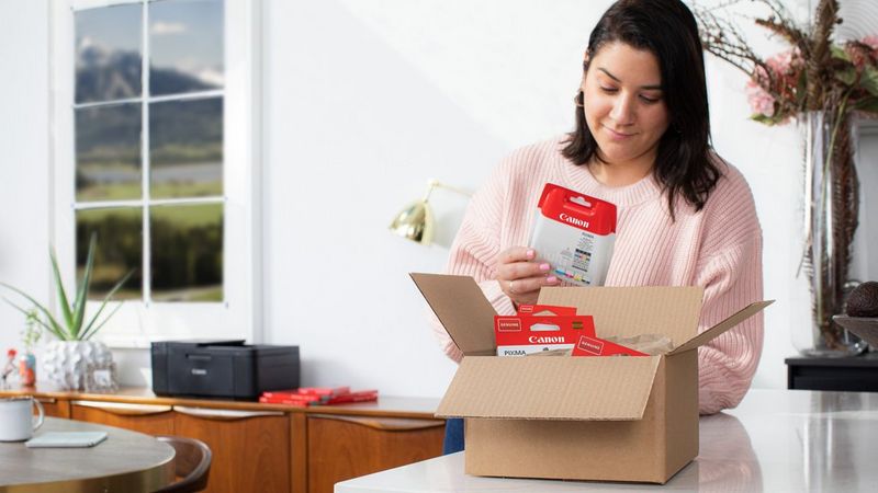 A user looks at a packaged ink cartridge, with others in a cardboard box in front of her and a Canon printer in the background.