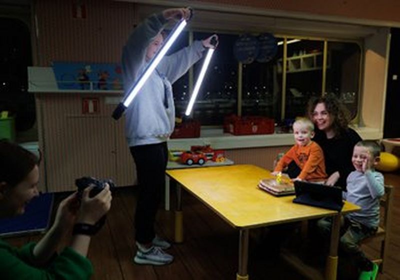 A teenaged girl holds LED light tubes in front of a woman and two small children who are sat at a table. Behind her, a woman holds a camera to photograph them.