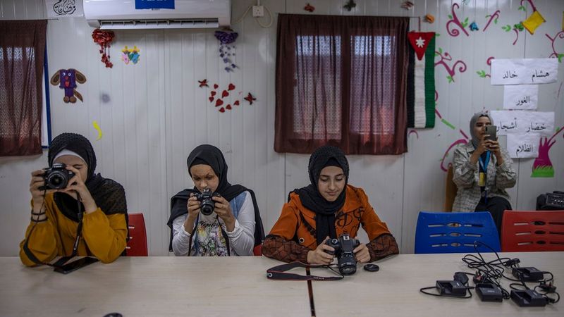 Three young women in black headscarves sit at a table, each is inspecting the back of a Canon camera, either by holding it and looking at the display or looking through the viewfinder. To the right, stood behind the table, another girl holds up a mobile phone, as though taking a picture or filming. They are in some kind of classroom, with drawings, colourful cutouts and cartoons stuck to the wall behind them.