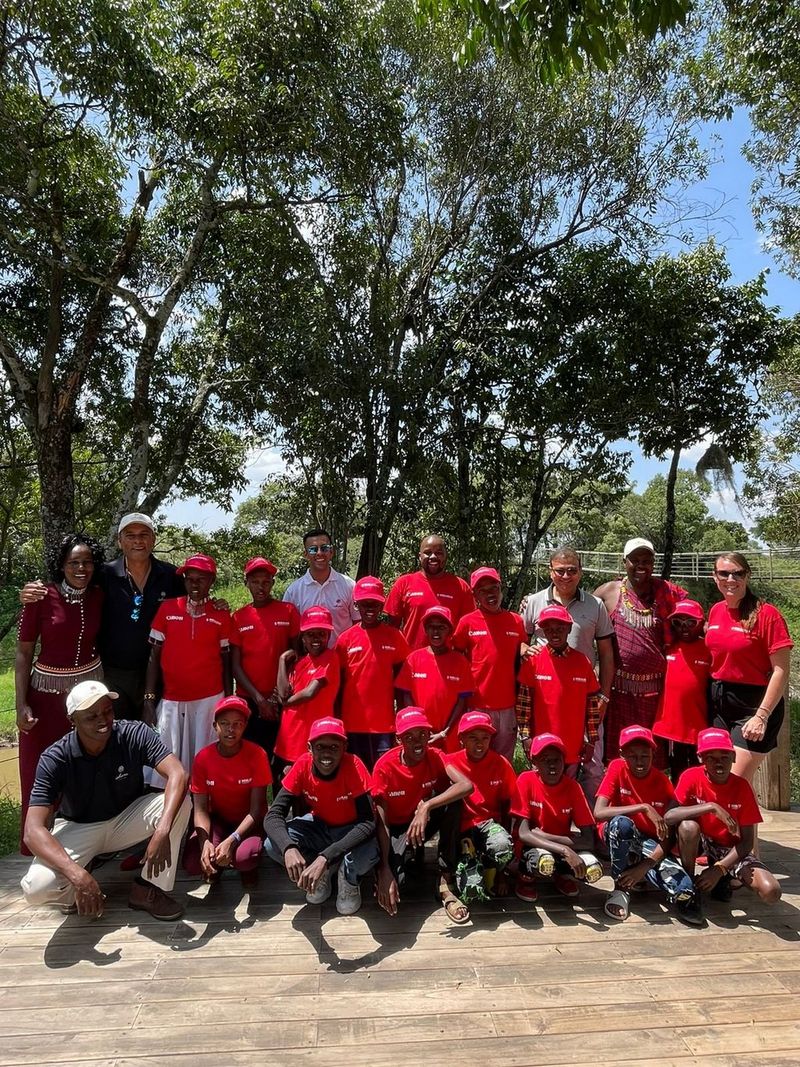 23 people, most in red Canon branded t-shirts gather for the camera in front of trees on a sunny day.