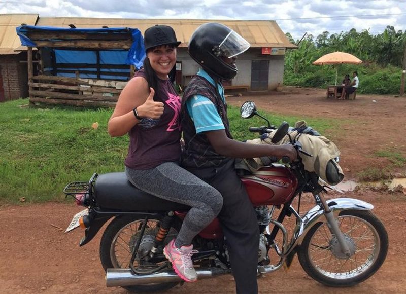 A woman rides pillion on a motorbike. She grins for the camera and gives the thumbs up.