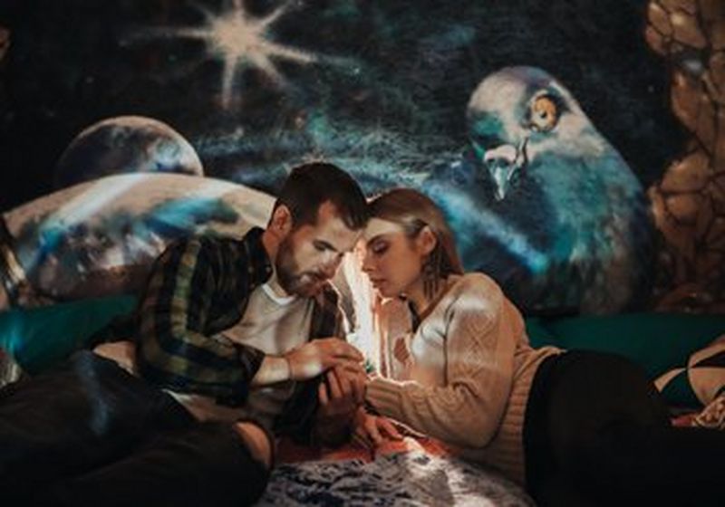 A couple, lying on blankets and cushions. holding hands, with their heads close together, looking down at their hands. In the background are images of birds and nature. © Katya Mukhina shot on Canon EOS R + Canon RF 28-70 f/2 L USM