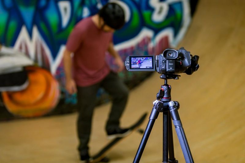 A man performing skateboard stunts is filmed with a Canon LEGRIA GX10 camcorder attached to a tripod.