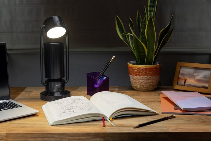 A Canon Light & Speaker ML-A is positioned on a wooden desk, next to a book, a plant and some stationery.