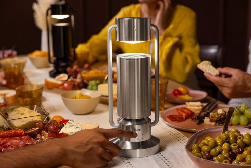 Two Canon Light & Speaker ML-As sit on a crowded dinner table, surrounded by plates of food and people sitting on chairs. A hand is reaching out and adjusting the controls on the nearest Light & Speaker ML-A.