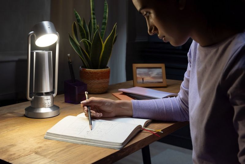 A Canon Light & Speaker ML-A is positioned on a wooden desk next to a woman working, illuminating the notebook she is reading from.