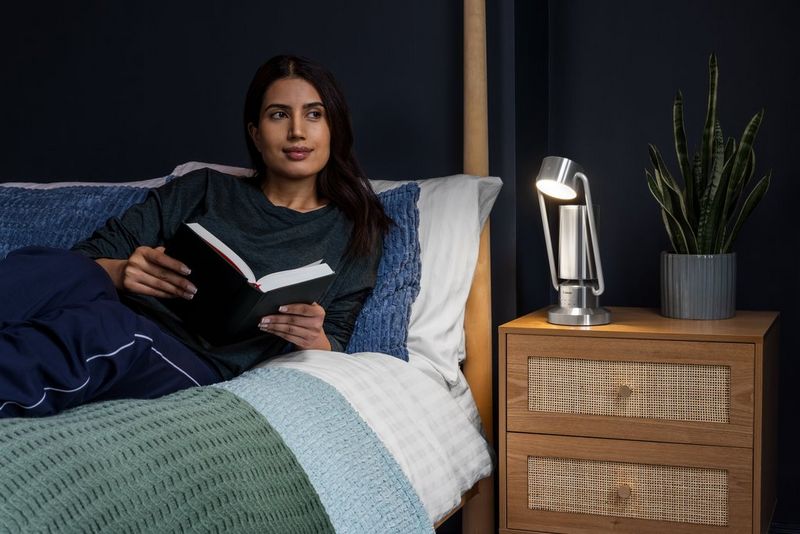 A person reclines in bed with a book, a Canon Light & Speaker ML-A on their bedside table.