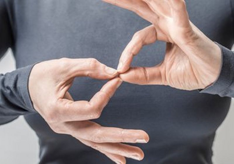 The hands, arms and torso of a woman, who is wearing a grey long-sleeved top. Her hands are making the British Sign Language sign for ‘linguistics’, the thumb and forefinger of each hand forming a circle and meeting at the fingertips.