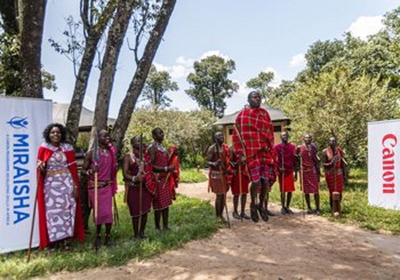 Ten men and women wearing the traditional dress of the Maasai tribe stand on a grassy verge between two pop-up banners displaying the Miraisha and Canon logos. The man in the centre of the image jumps high into the air. © Peter Ndungu 