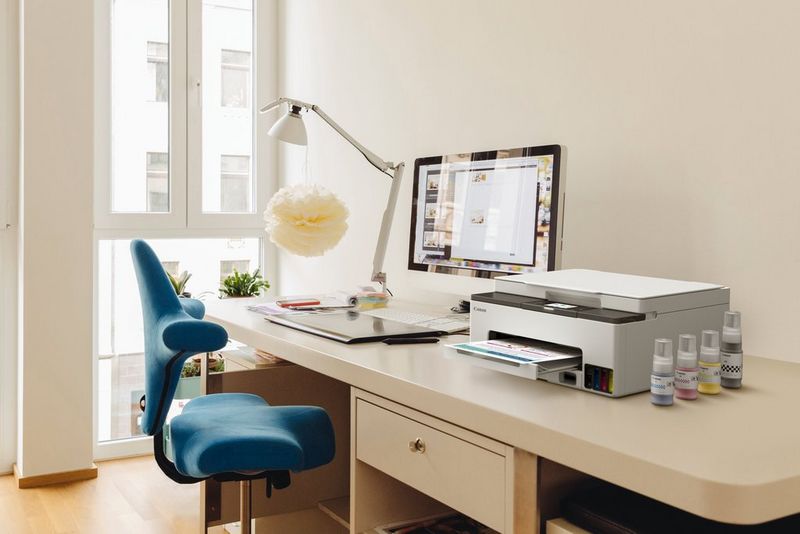 A Canon MegaTank printer and some bottles of replacement ink sit on a desk with a computer monitor and desk lamp behind. 