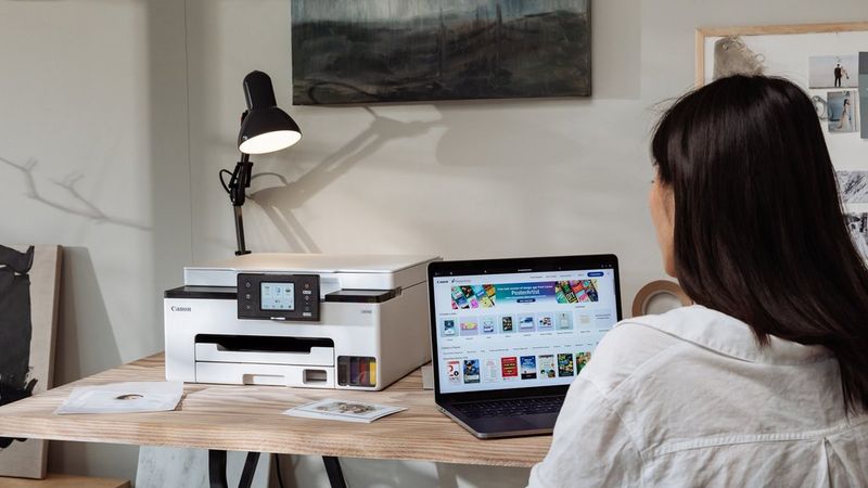 A woman sits working at a laptop, with a Canon MegaTank printer on the desk next to her.