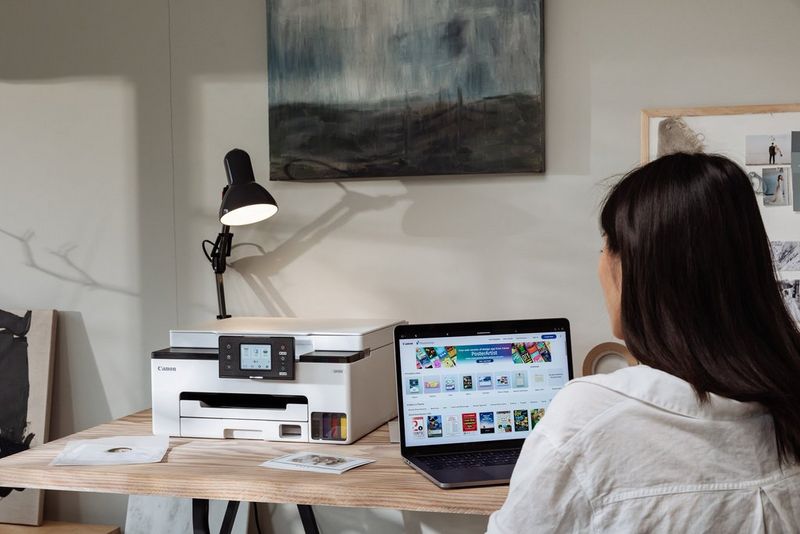 A person sits at a small desk working on a laptop. On the desk behind the laptop is a Canon PIXMA printer.