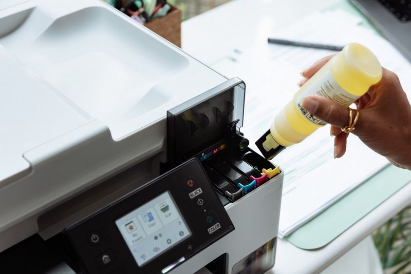 A user's hand holds a bottle of yellow ink ready to insert into a Canon MegaTank printer. 