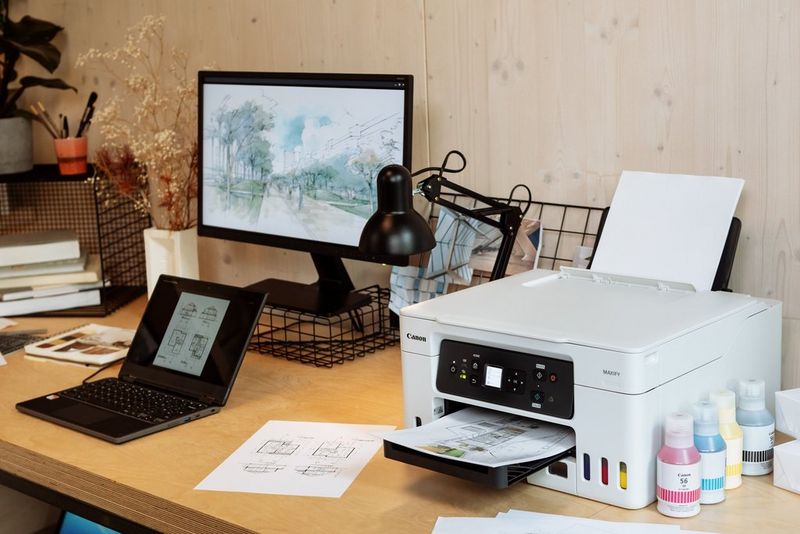 On a desk, a Canon MAXIFY printer sits next to a lamp, laptop, screen, plant and stationery. 