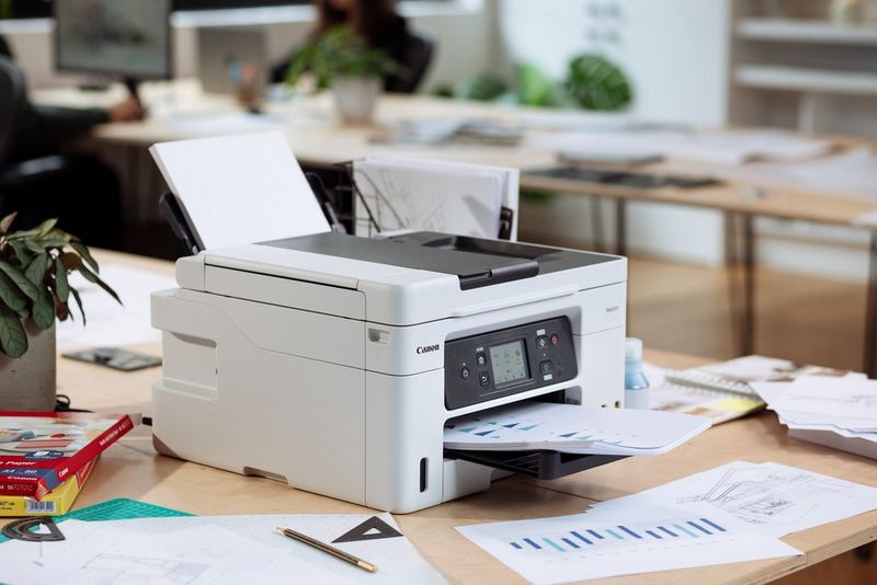 A Canon MAXIFY printer sits next to scattered papers on a table. 