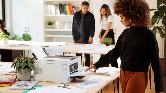 A woman picking up a printout from a Canon MAXIFY printer in an office with two other people in the background.