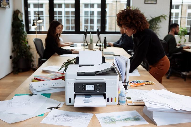 A print emerges from a Canon MAXIFY printer on an office desk with piles of papers around it. Five people sit at desks in the background.