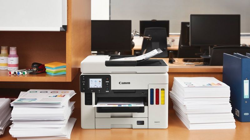 A MAXIFY printer on an office desk surrounded by piles of printouts.