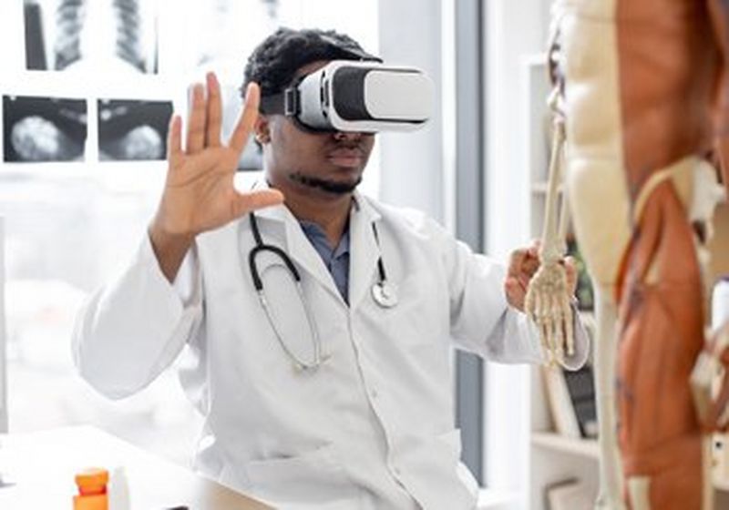 A doctor in a white coat and wearing a VR headset holds his hands up. Beside him is an anatomical skeleton and some scans are stuck to a board behind him.