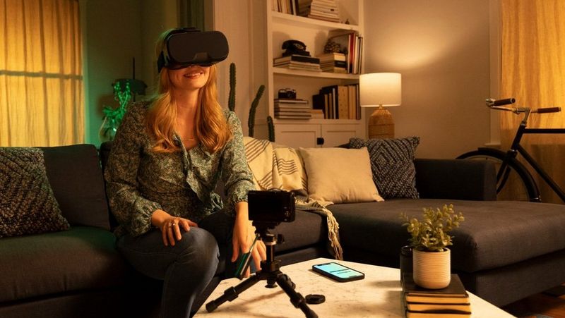 Women sits in living room on sofa wearing VR headset. A small camera and mobile phone are in front of her on a coffee table.