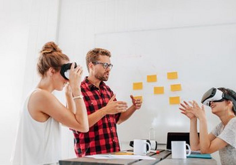 Three casually dressed people in a white-walled room sit and stand around a wooden table. Two are wearing VR headsets and a third headset sits on the table, alongside white coffee mugs, pens, paper, a notebook and a laptop. On the wall next to them are seven yellow sticky notes.