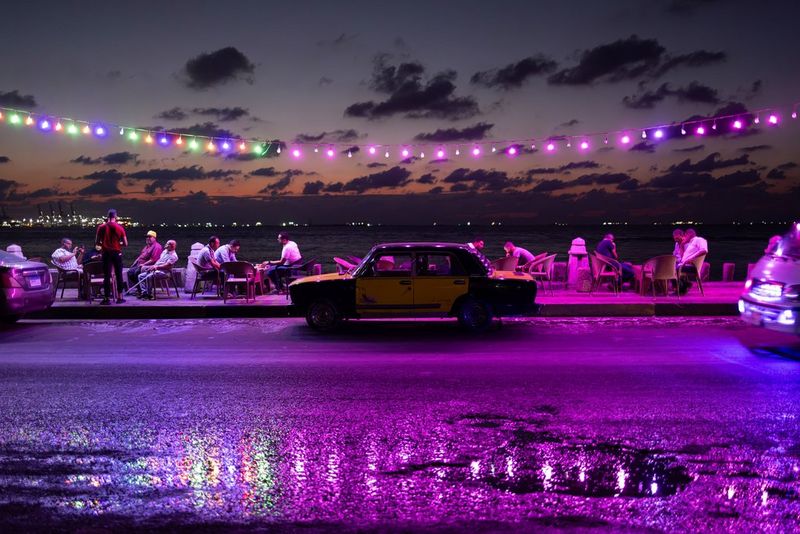 Varios grupos de personas se sientan juntos con una playa a lo lejos; varios coches aparcados ocupan la carretera mientras unas guirnaldas de luces violeta se reflejan por encima. Imagen tomada con una Canon.