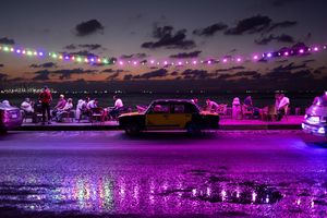 Gruppi di persone sedute insieme davanti a una spiaggia, auto parcheggiate lungo la strada e lucine viola che si riflettono al di sopra. Scatto realizzato con Canon.