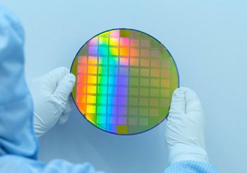 A person in a cleanroom suit holds a silicon wafer up in two gloved hands.