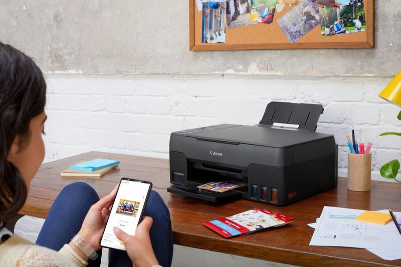 A young woman holding a smartphone sits at a desk with a Canon PIXMA printer on it. 