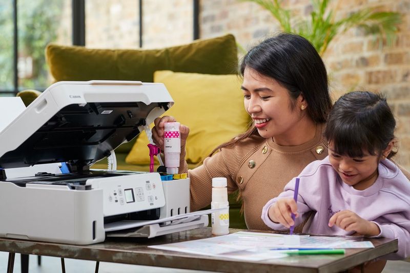 A smiling woman and child sit at a table alongside a Canon PIXMA MegaTank printer. The woman is refilling the printer's ink tanks while the child is colouring in a printout.