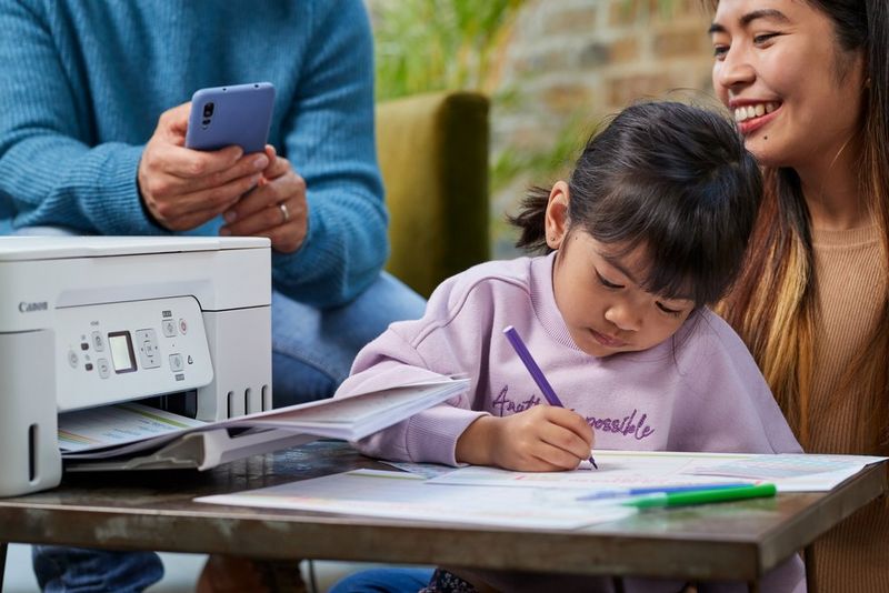A child sits on a smiling woman's knee colouring in printouts from a Canon PIXMA printer. Hands holding a smartphone can be seen in the background.