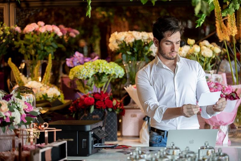 A man looks at a card in his hand, with a display of colourful flowers arrayed behind him and a Canon printer to one side.