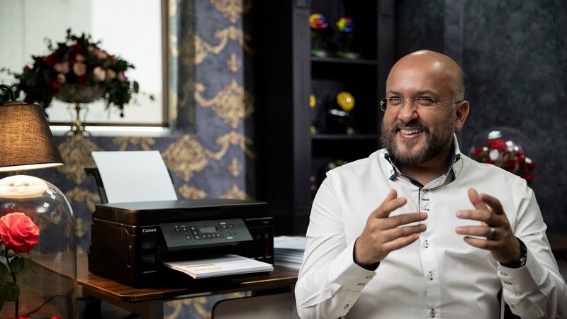 A man sitting, talking, and smiling next to a Canon PIXMA printer on a table.