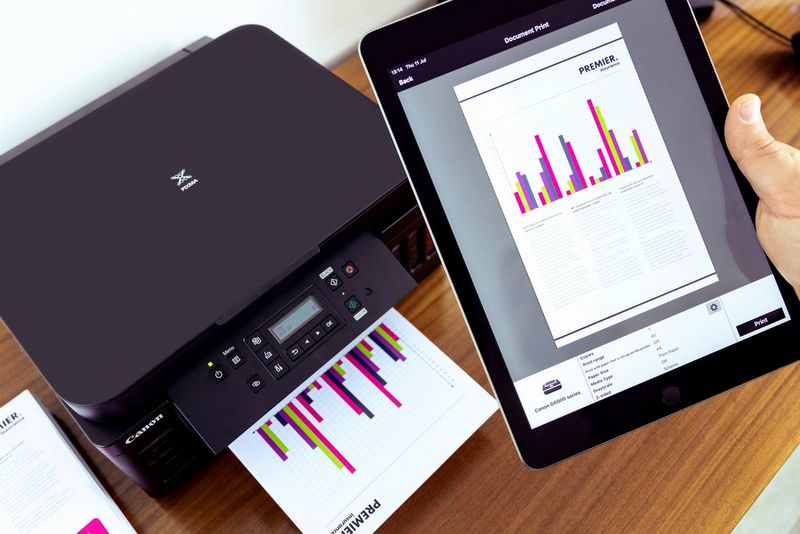 A user holds a tablet with a bar chart on the screen while a print of the same chart emerges from a Canon printer.