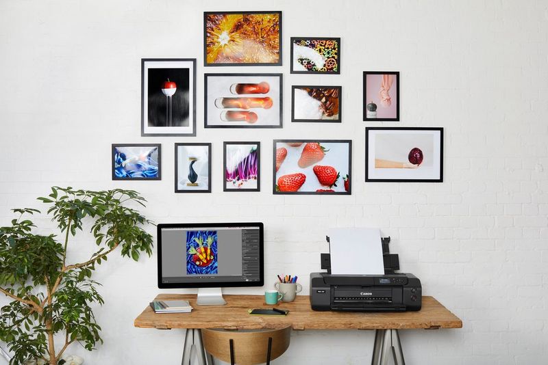 A Canon printer and monitor on a wooden desk. On the wall above are 11 colourful framed prints featuring food. 