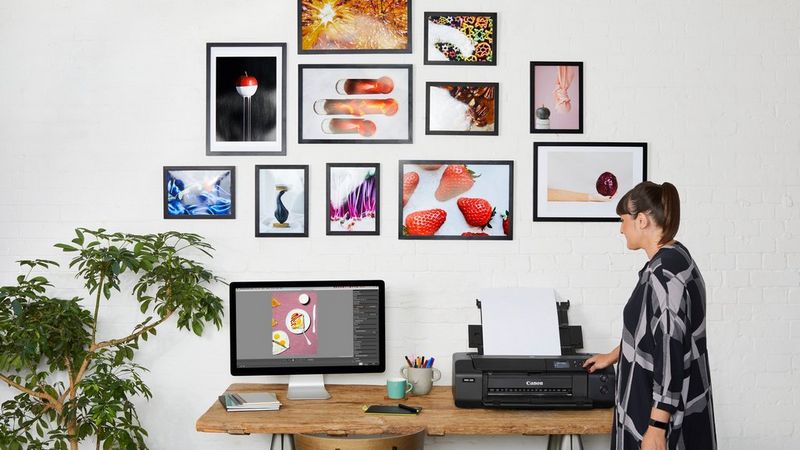 A woman standing by a Canon PIXMA printer, which is sitting on a desk next to a computer monitor. Printed photos are displayed in frames on the wall behind.