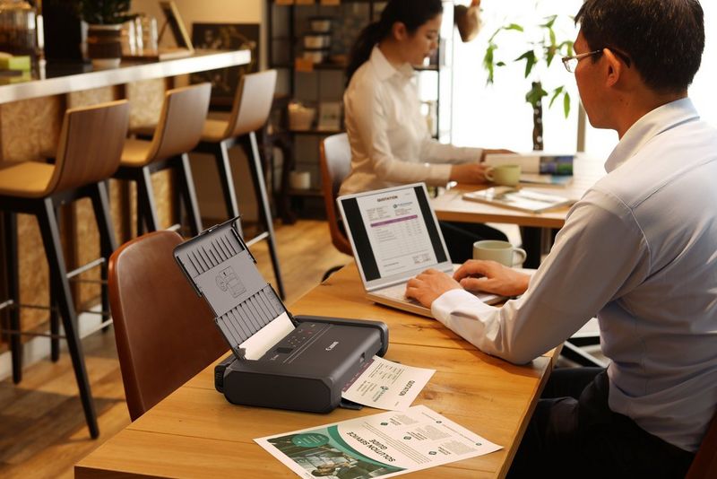 A man sitting in a cafe with a laptop prints documents on a Canon PIXMA TR150 printer.