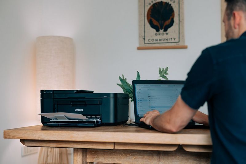 A man sits at a table working at a laptop. Next to it is a Canon printer with a sheet of paper in the output tray.