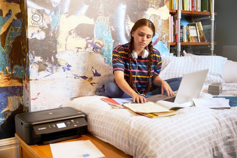 A teenager sits cross-legged on a bed, studying textbooks. A Canon PIXMA printer is on the table beside the bed.