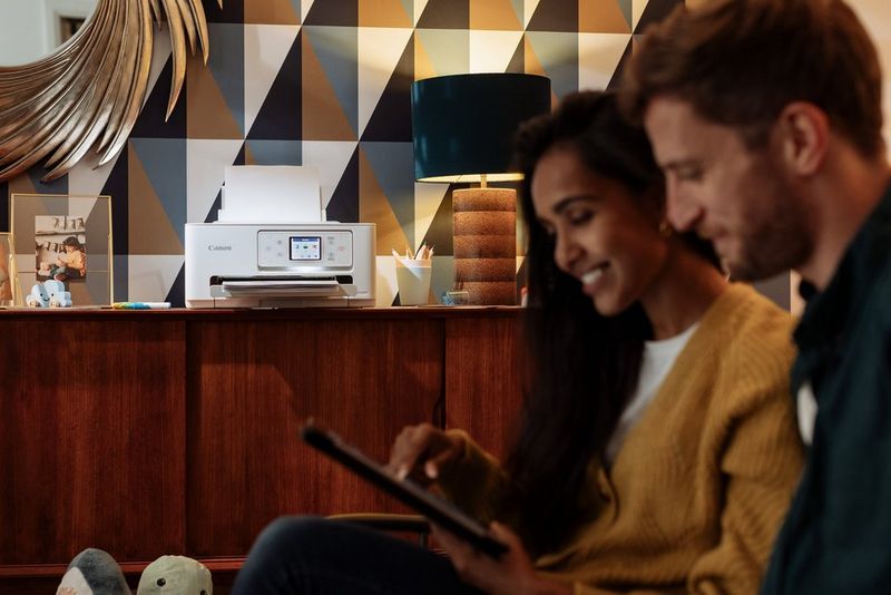 A couple sit on a couch looking at phones and tablets. A PIXMA printer is on a sideboard behind them. 