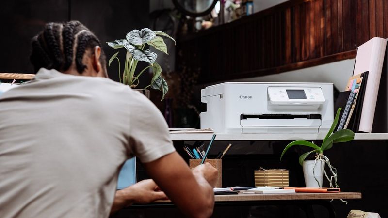 A young man with his back to the camera leans over a desk on which sits a Canon PIXMA printer, plants and stationery.
