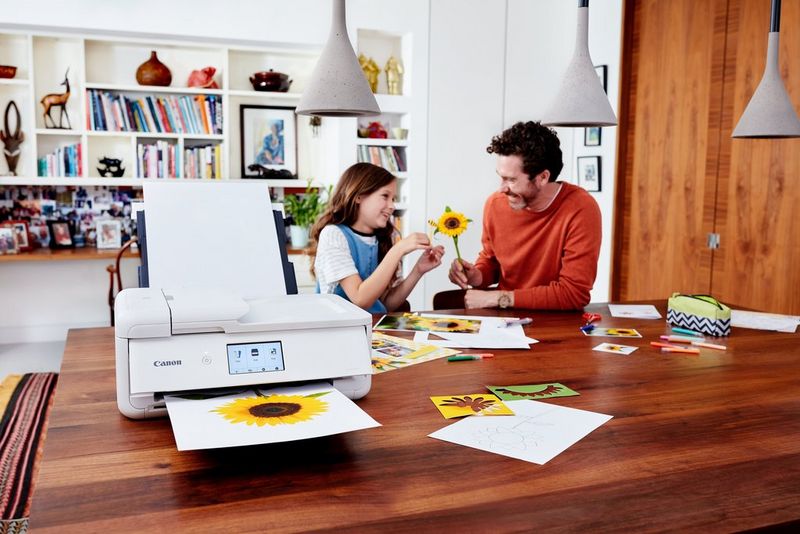 A Canon PIXMA printer on a table prints a template of a sunflower, while in the background a father and daughter are making the same sunflower.