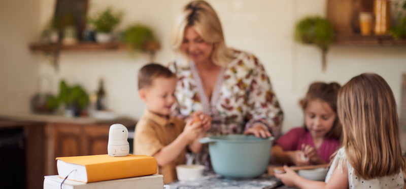 A woman and her three children standing around a mixing bowl in a kitchen as they prepare to bake something. The PowerShot PX sits on top of a pile of books facing them.