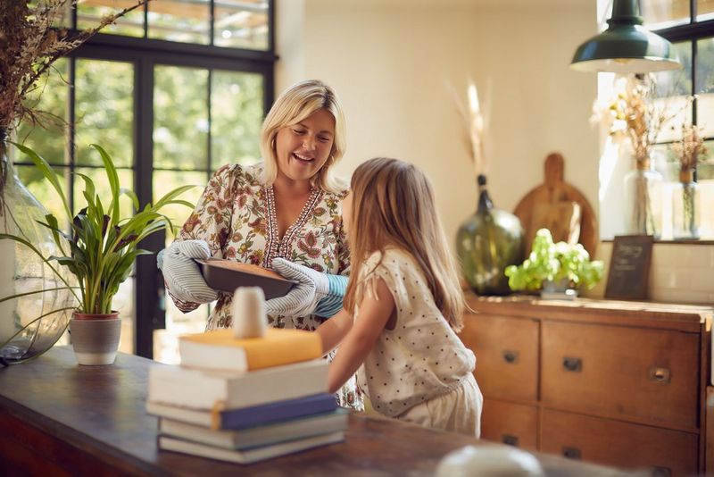 A mother smiles at her daughter as she takes a cake out of the oven. Blurred in the foreground, capturing the moment, is a Canon PowerShot PX.