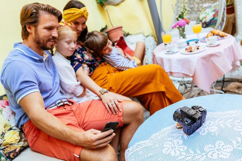 A group of adults and children sit together at a table, and the man on the left uses his smartphone to trigger the shutter on the Canon camera on the table in the foreground.