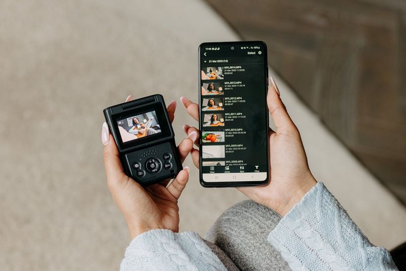 A user holds a Canon PowerShot V10 in one hand and a smartphone with the Canon Camera Connect app transferring files from the camera in the other.