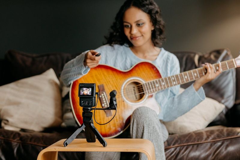 A woman playing the guitar looks into a Canon PowerShot V10 with an external microphone and Canon Tripod Grip HG-100TBR attached, standing on a low table facing her.